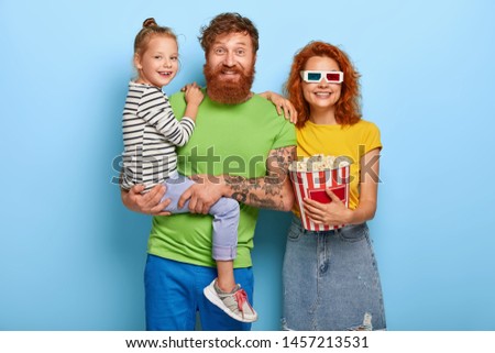 Merry ginger family have friendly relationship, spend free time all together, husband and wife visit movie with daughter, watch cartoon, woman wears three dimensional glasses, holds popcorn.