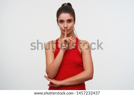 Portrait of young mysterious cute brunette woman wears in red t-shirt shows silence gesture, please keep calm. Stands over white background.