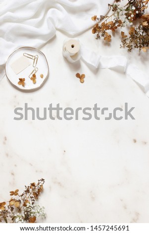 Wedding, birthday stationery styled stock photo. White table runner, porcelain plate with stamp, golden clips, silk ribbon, dry hydrangea, gypsophila flowers. Marble stone background. Vertical flatlay