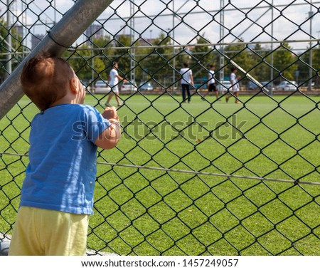 Baby boy looking at children playing football. A cute 1 year old baby boy watching a soccer or a football game near the field behind the metal wire fence. Sport and football fan from babyhood.