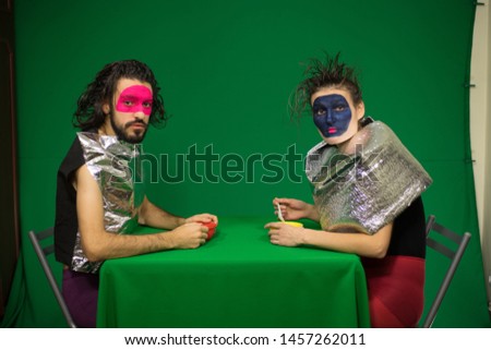 Couple sitting at the green table. Green background. Actors in the studio. Clowns. Futuristic makeup. Futuristic costumes.