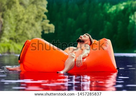 A young man is resting lying in an inflatable orange lounger floating on the water surface of a forest river. Camping during eco-tourism.