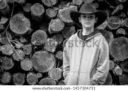 Black and white portrait of a young cowboy wearing a dark hat. Behind him, a pile of logs