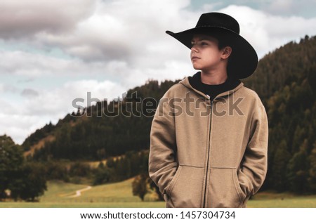 Young cowboy wearing a black hat. Behind him, a mountain landscape