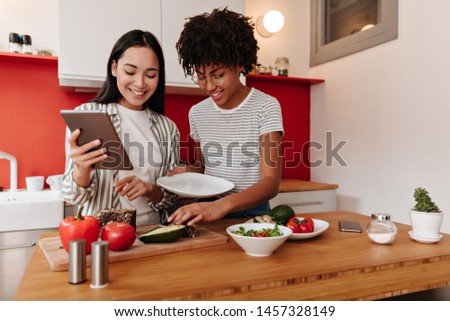 Women in T-shirts with smile look at tablet and lay vegetables on plate