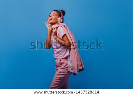 Lovely relaxed woman in headphones singing on blue background. Carefree black girl laughing during photoshoot.