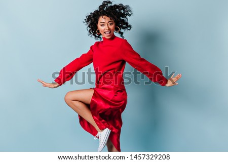 Positive girl with wavy hair dancing on blue background. Studio shot of happy young woman in red clothes.