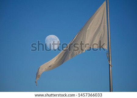 White flag flying against blue sky with large visible moon. Sun is setting, and gives the flag a faint glow. Pure blue and white two tone image. Isolated flag, big moon, and flag pole visible.