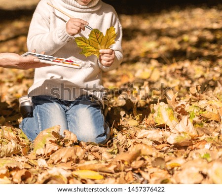 Cute little girl  sitting and drawing with paint autumn leaves outside. Park and Autumn Leaves on a Background. Autumn season concept.