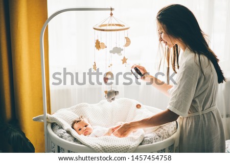 Mother taking a picture of A baby with a mobile phone. Mother holding daughter hand and smiling. Mother standing near the cradle and holding baby's hand. Concept photo parenthood and motherhood.