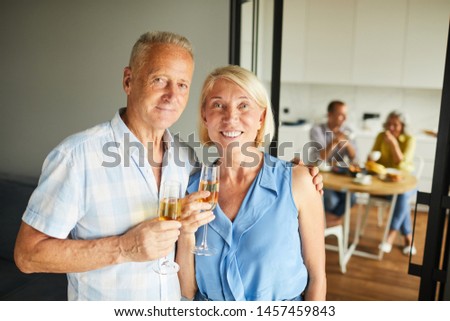 Waist up portrait of smiling mature couple looking at camera posing at home with friends in background, copy space
