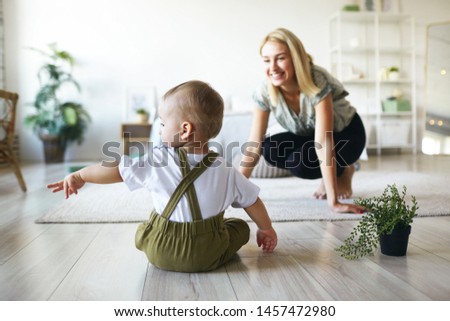 Back view of infant child in stylish clothes sitting on floor with plant pot, raising hand, pointing finger sideways, showing something to his young cheerful mother. Family, motherhood and infancy