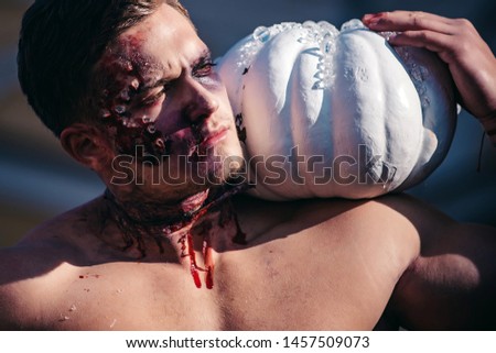 Muscular man with broken head. Close-up portrait of a man with zombie makeup. Scary and bloody muscular zombie man. Young handsome man with muscular body posing in street holding halloween pumpkin