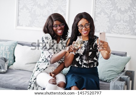 Two african woman friends wear on eyeglasses posed indoor white room, drinking champagne and making selfie.
