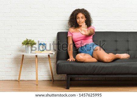 Young african american woman sitting on the sofa standing with outstretched hand showing stop sign, preventing you.