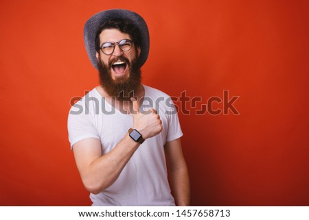 Photo of excited bearded guy in hat and glasses, showing thumb up over red backgroung 