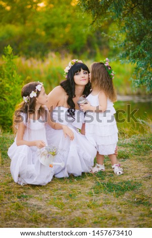 Young mother with her daughters in white dresses having fun in a summer field at sunset by the river