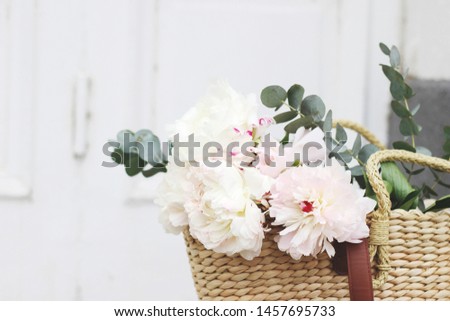 Feminine wedding still life composition. Straw French basket bag with pink peonies flowers and eucalyptus bouquet. Old white door in the background. Styled stock photo. Selective focus.