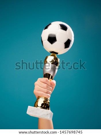 Photo of hand holding victory cup with soccer ball on empty blue background