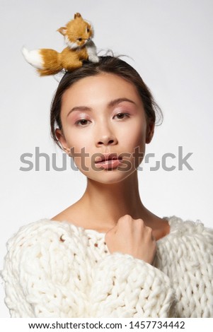 Cropped half-turn shot of upper body’s part of a lady, wearing white knitted sweater and headband with figurine of a fox. 