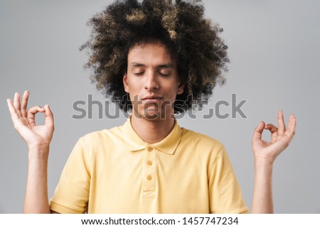 Photo of calm caucasian man with afro hairstyle meditating and keeping fingers in zen gesture isolated over gray background