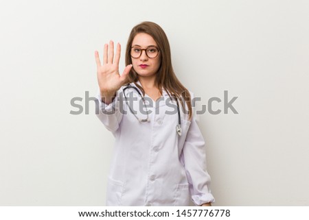 Young doctor woman against a white wall standing with outstretched hand showing stop sign, preventing you.