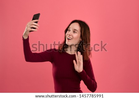Photo of a pretty young brunette woman making selfie with a mobile phone holding hands in a gesture of Hi