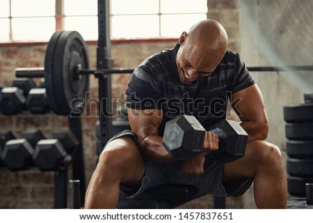 Muscular guy lifting dumbbell while sitting on bench at gym. Mature african athlete using dumbbell during workout. Strong man under physical exertion pumping up bicep muscle with heavy weight. Royalty-Free Stock Photo #1457837651