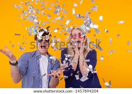 Young cheerful positive couple of funny glasses and a paper cap rejoice and shout on a yellow background with flying confetti. Conception of a holiday and fun.
