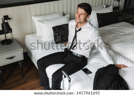 Image of pleased young man wearing formal suit sitting on bed with suitcase in hotel room during business trip