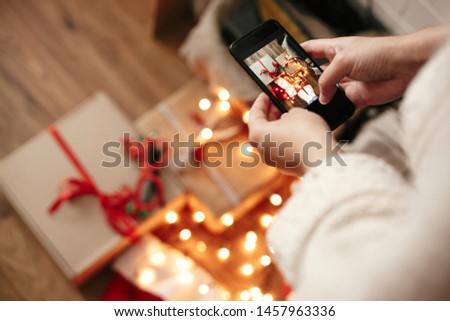 Hands holding phone and taking photo of christmas gift boxes, santa hat, illumination lights on wooden background in dark room. Stylish hipster girl in sweater making christmas flat lay photo