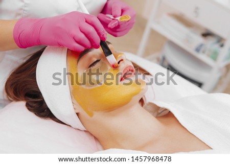 Hands of cosmetology specialist applying gold facial mask using brush, making skin hydrated and face glowing and skin. Attractive brunette relaxing with closed eyes and enjoying spa procedures. Royalty-Free Stock Photo #1457968478