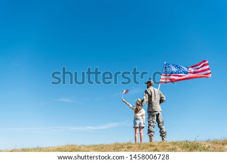 back view of child in straw hat and military father holding american flags while standing on grass 