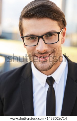 Close up of a handsome young businessman wearing suit standing outdoors at the city streets, looking at camera