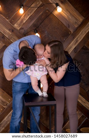 Happy Young Caucasian Family in Studio. Mother and Father Standing near Chair. Parents Kissing Little Daughter. Baby Standing on Chair. Infant Holding Pink Flower. Wooden Background with Lightbulbs.