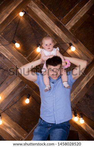 Happy Young Caucasian Family in Studio. Father Sitting on Chair. Man Holding Little Daughter on His Neck. Infant Holding Pink Flower. Funny Child. Wooden Background with Lightbulbs.