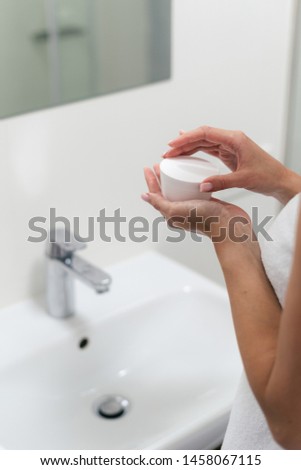 Cropped view and vertical photo of woman standing in bright light bathroom near mirror, holding cosmetics jar with face cream in hands