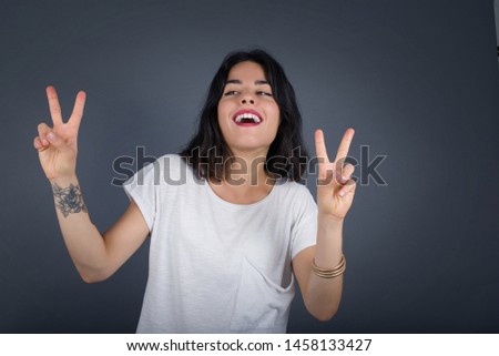 Isolated shot of cheerful European woman makes peace or victory sign with both hands, dressed in casual clothes, feels cool has toothy smile, isolated over gray background. People and body language.