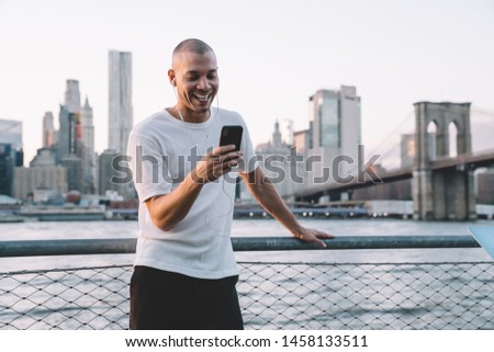 Young laughing ethnic man with earphones using smartphone while leaning on metallic fence with chain link against background of bridge and city