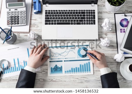 Businessman working with financial reports. Flat lay workplace with man hands, laptop and paper documents on wooden desk. Business presentation with diagrams. Investment and stock market analysis.