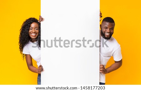 Black Girl And Guy Holding White Advertising Board, yellow studio background, panorama Royalty-Free Stock Photo #1458177203