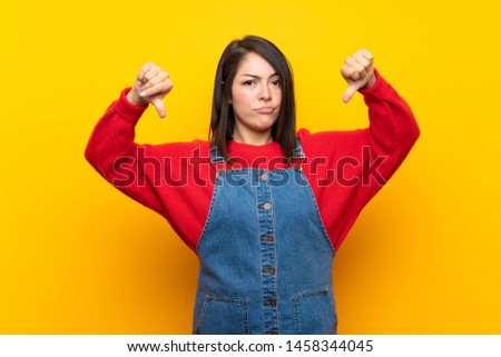 Young Mexican woman with overalls over yellow wall showing thumb down