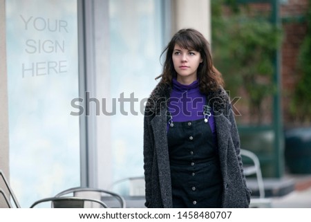 Woman walking outdoors in the city looking at text or logo space on a window display.  Advertising or promotional graphics can be added to the blank wall outside the store.  She is window shopping. 