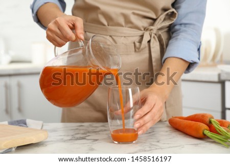 Woman pouring tasty carrot juice from jug into glass at table indoors, closeup