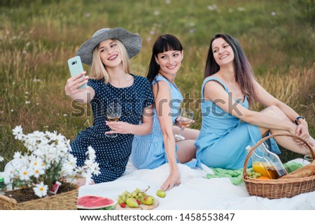 Three young women, in blue dresses and hats sit on plaid and take pictures on smartphone. Outdoor picnic on grass. Delicious food in picnic basket and wine. Watermelon, grapes and bouquet of daisies.