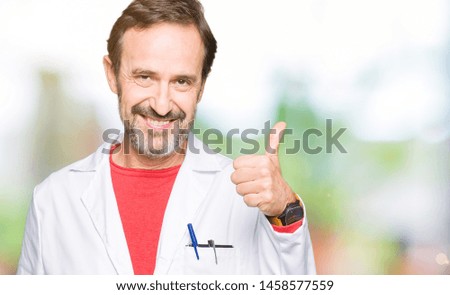 Middle age therapist wearing white coat doing happy thumbs up gesture with hand. Approving expression looking at the camera with showing success.