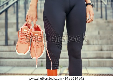 New day. Cropped photo of woman with leg prosthesis in sports clothing carrying her orange sneakers while standing on stairs outdoors