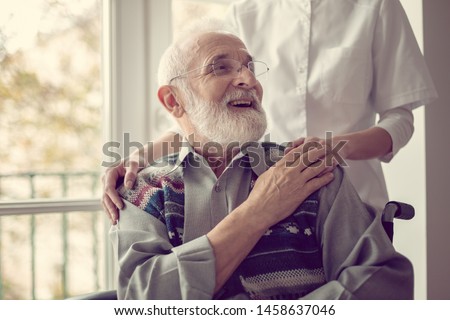 Senior man sitting on the wheelchair, laughing and holding his nurse's hand Royalty-Free Stock Photo #1458637046