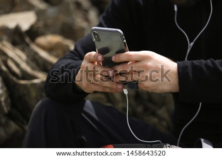 Close-up picture of black modern smartphone with white earphones. Man holding phone in hands and typing, searching in internet. Wearing black clothes. 