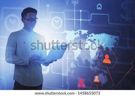 Smiling Asian man in glasses with laptop over blurred blue background with world map and social network hologram. Concept of hi tech and recruitment. Toned image